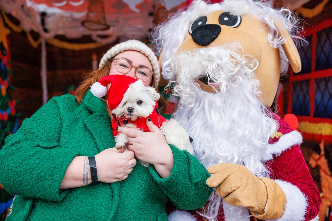 A woman holding a small white dog in a Christmas hat next to a person dressed as a dog Santa