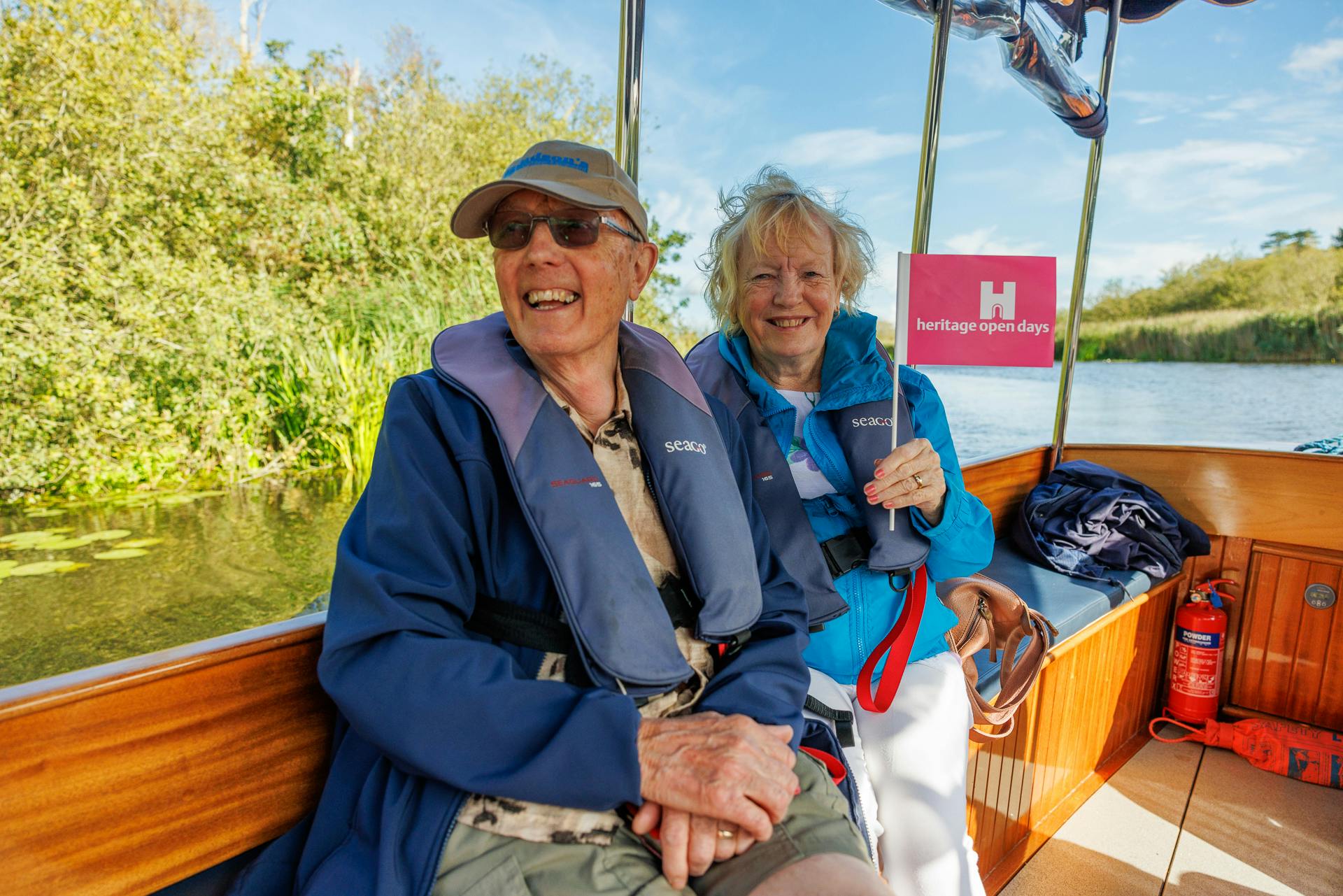 A man and a woman wearing lifejackets on a boat on the river smile to the camera.