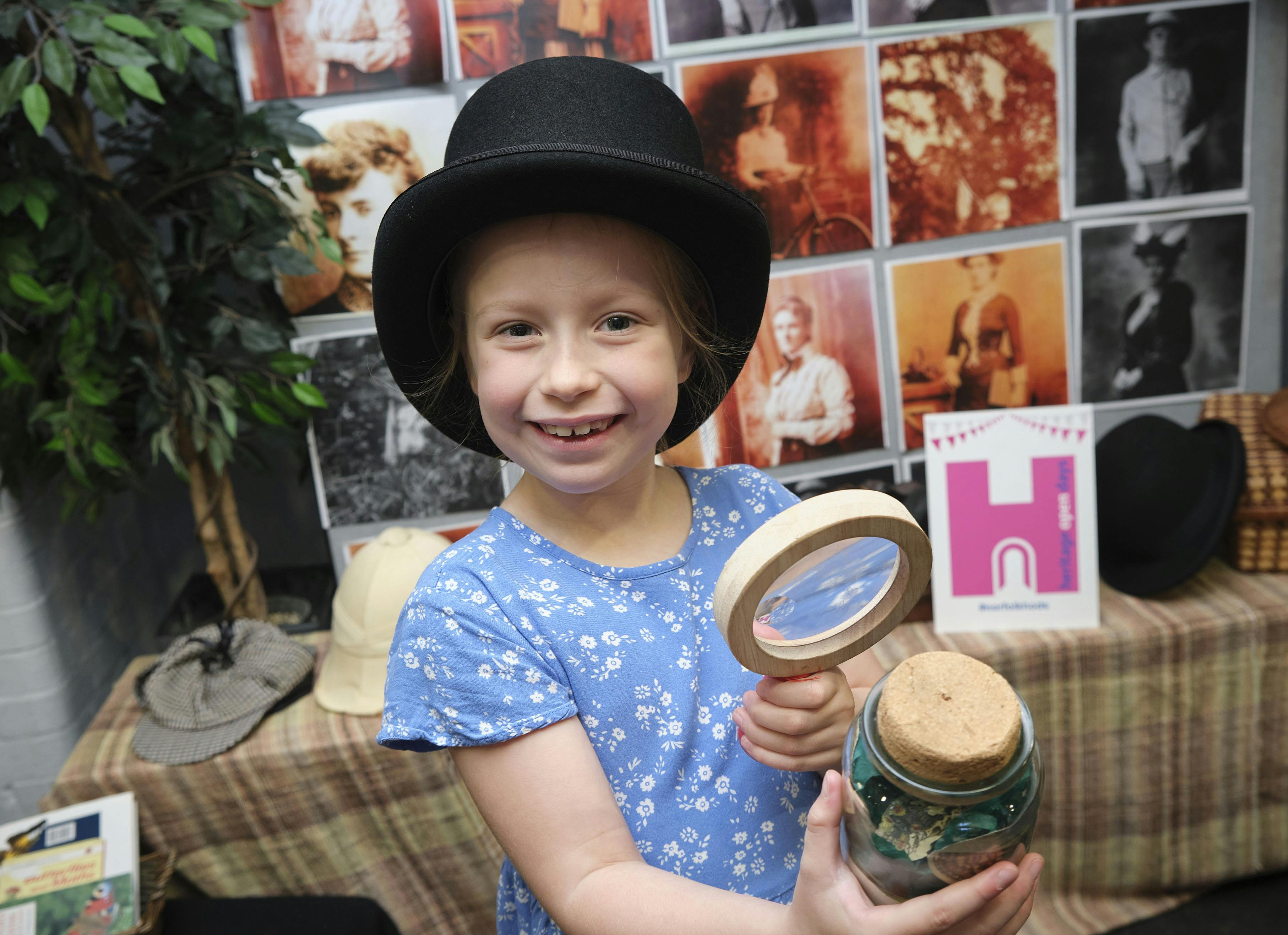A little girl wearing a blue dress and a top hat smiles to the camera