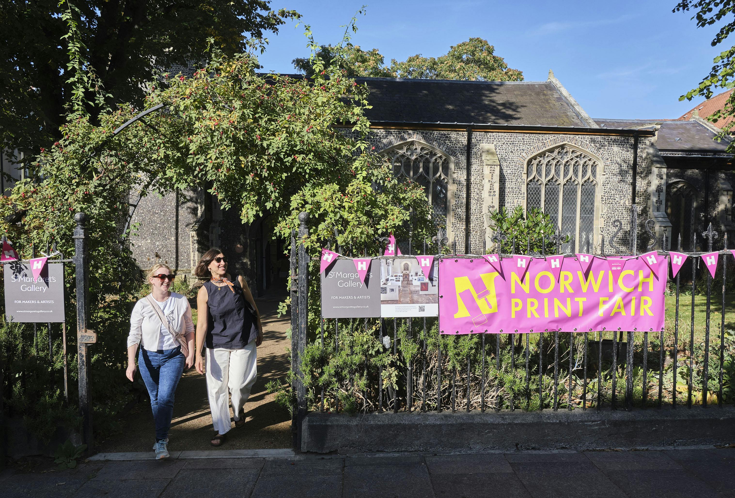 Two women smiling and walking out of a church garden with pink Heritage Open Days bunting on the iron church fence.