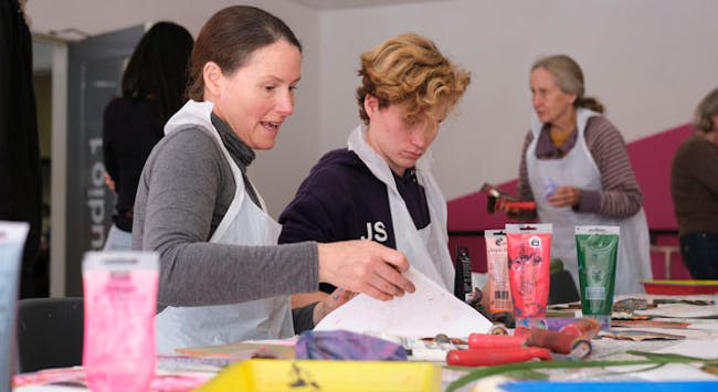 Two people with aprons on, making prints surrounded by ink bottles, rollers and paper.