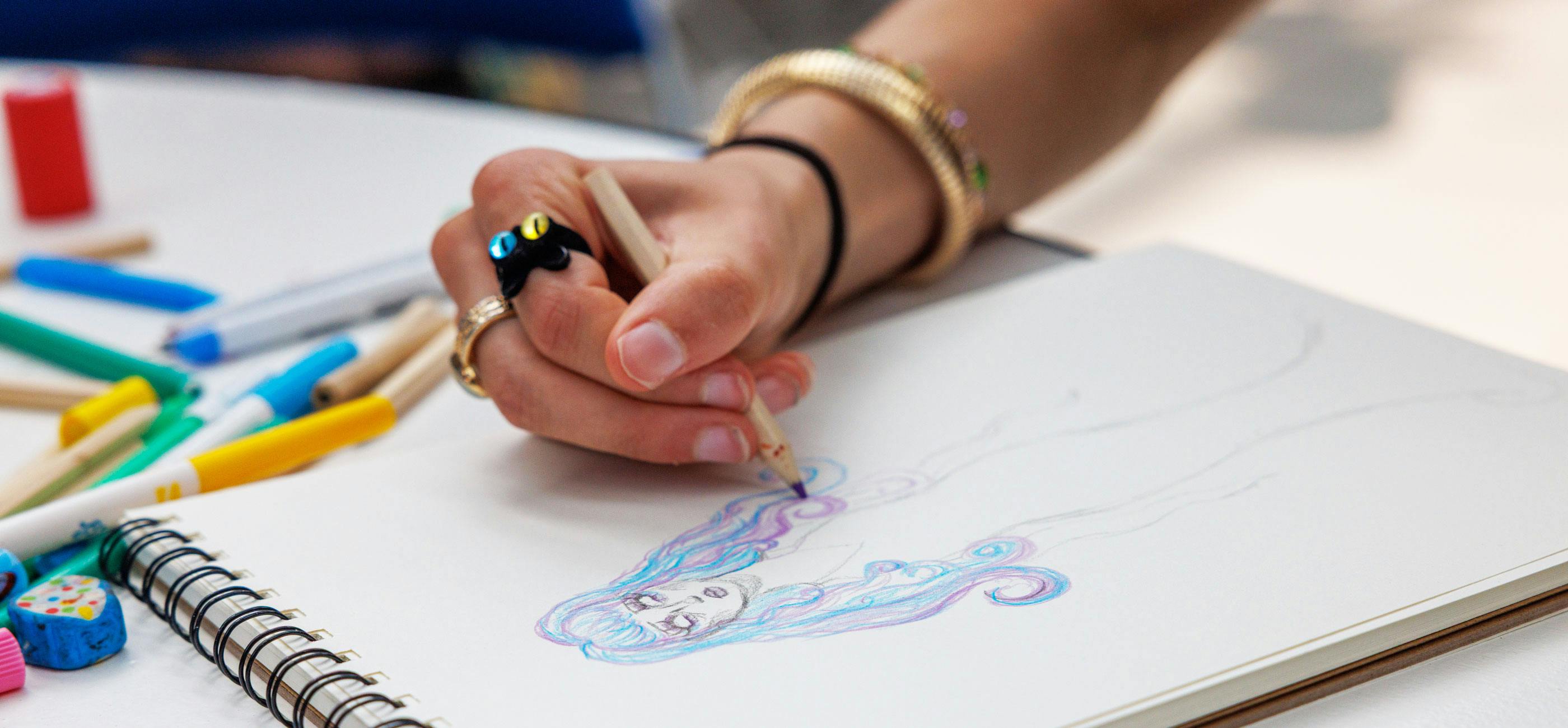 A close up of a hand drawing a colourful character with purple and blue hair