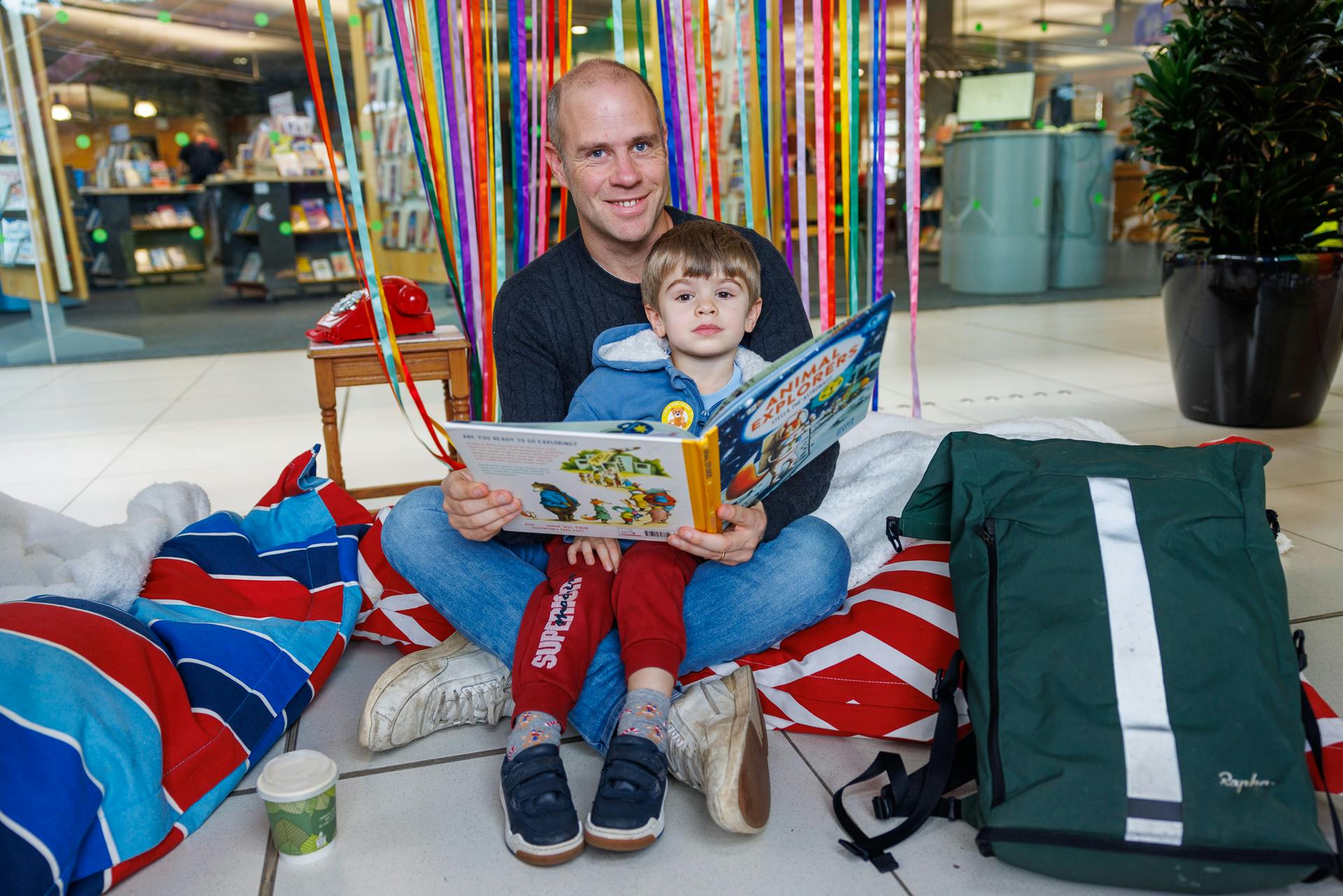 A father, sitting on cushions on the floor cross-legged with his young son sitting on his lap while reading a book called Animal Explorers, together