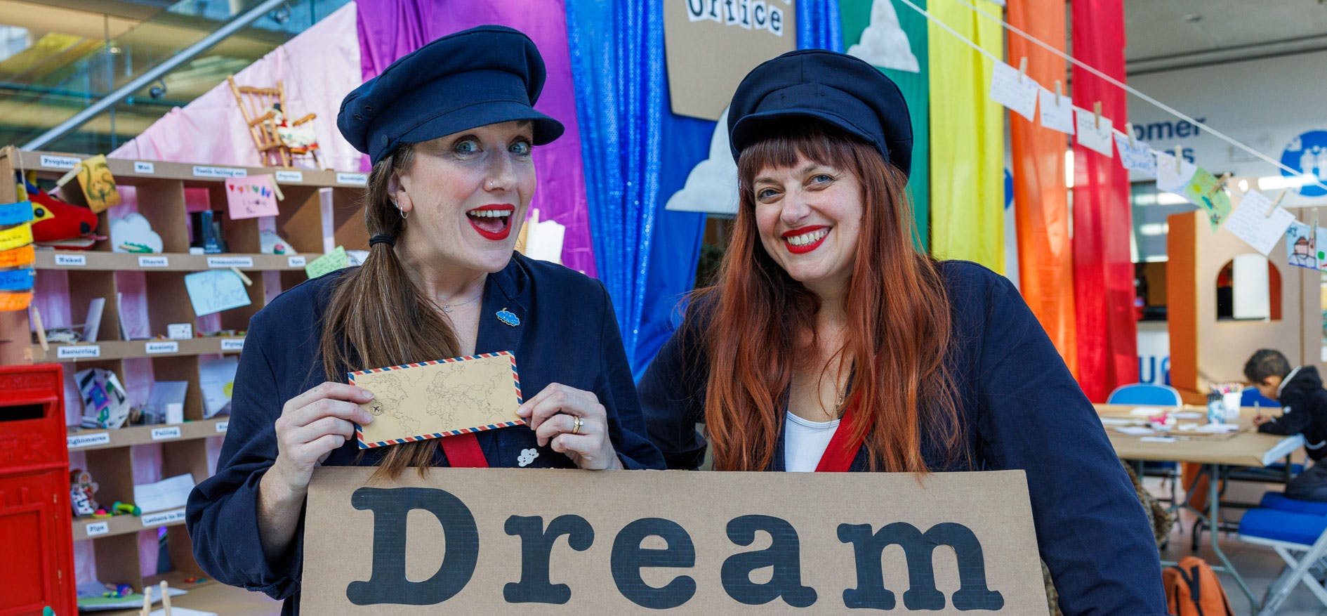 Two post women, in The Imaginarium sorting office with a sign saying Dream and rainbow colours in the background..