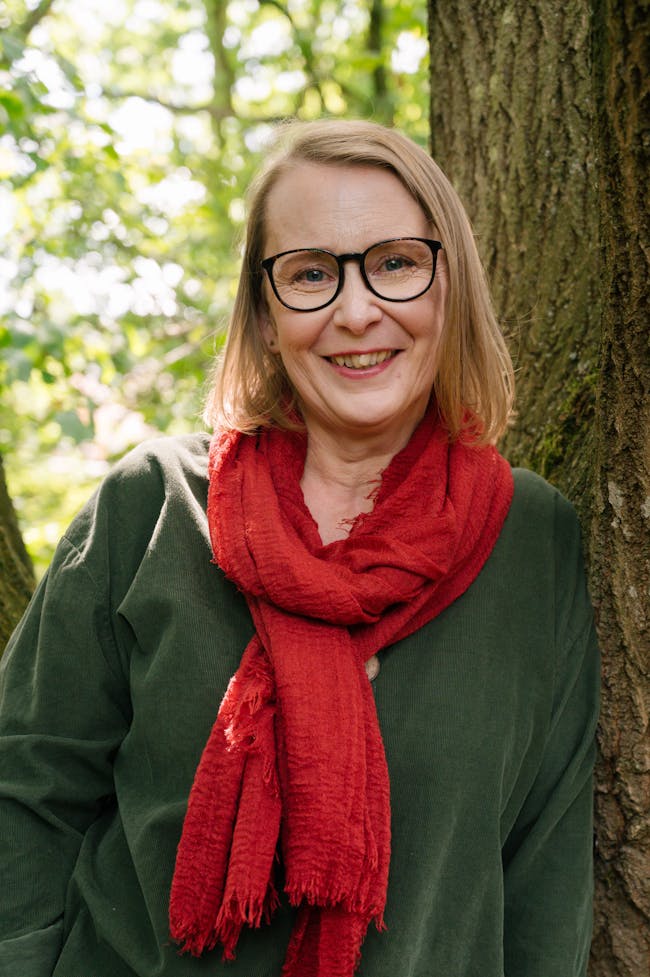 A head and shoulders shot of Heidi, who has mid-length blonde hair and glasses, leaning against a tree smiling at the camera.