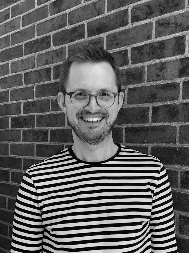 A black and white image of Matt Robertson, white man with glasses, in a stripey top in front of a brick wall. a stripey top