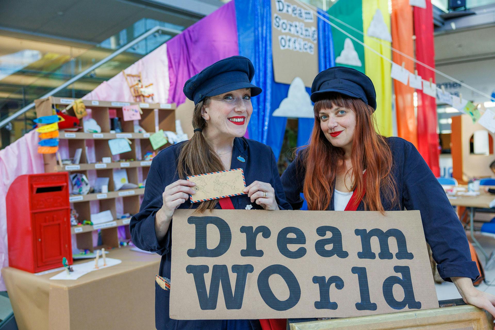 Two women dressed in post hats with rainbow background with the sign Dream World.
