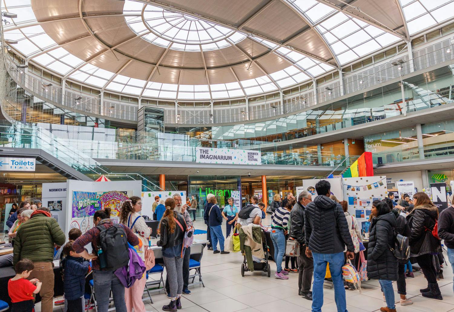 A busy book festival with lots of people at stands in a modern glass roofed building.