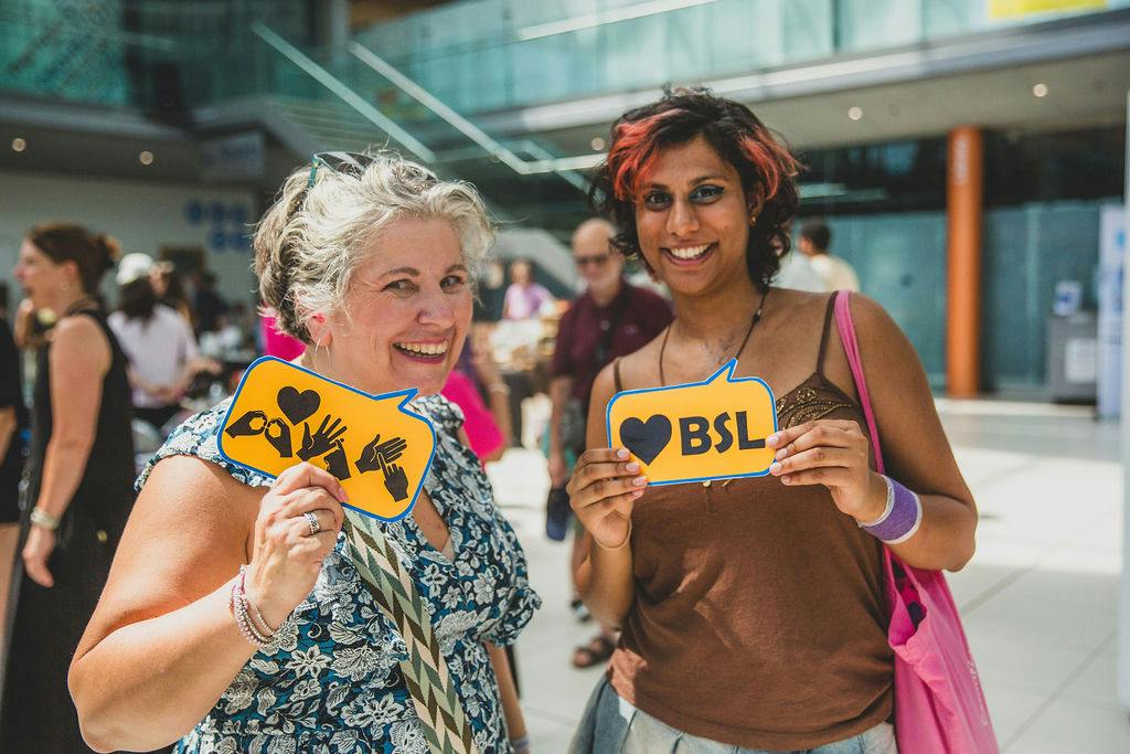 Two women in The Forum's Atrium, one white and one of colour, holding up BSL signs