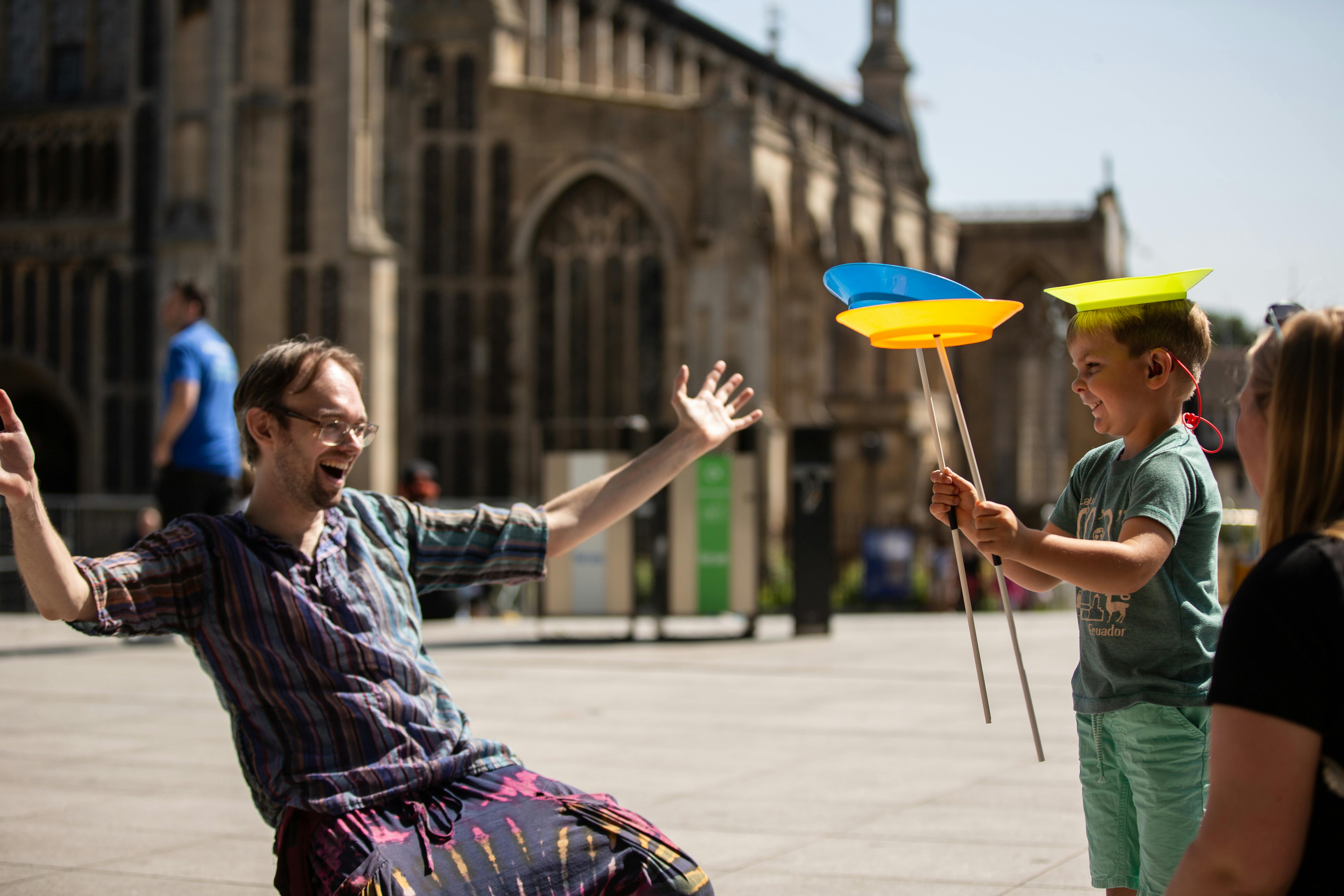 A child spinning two plates on sticks with one plate on his head, while a man cheers him on enthusiastically.