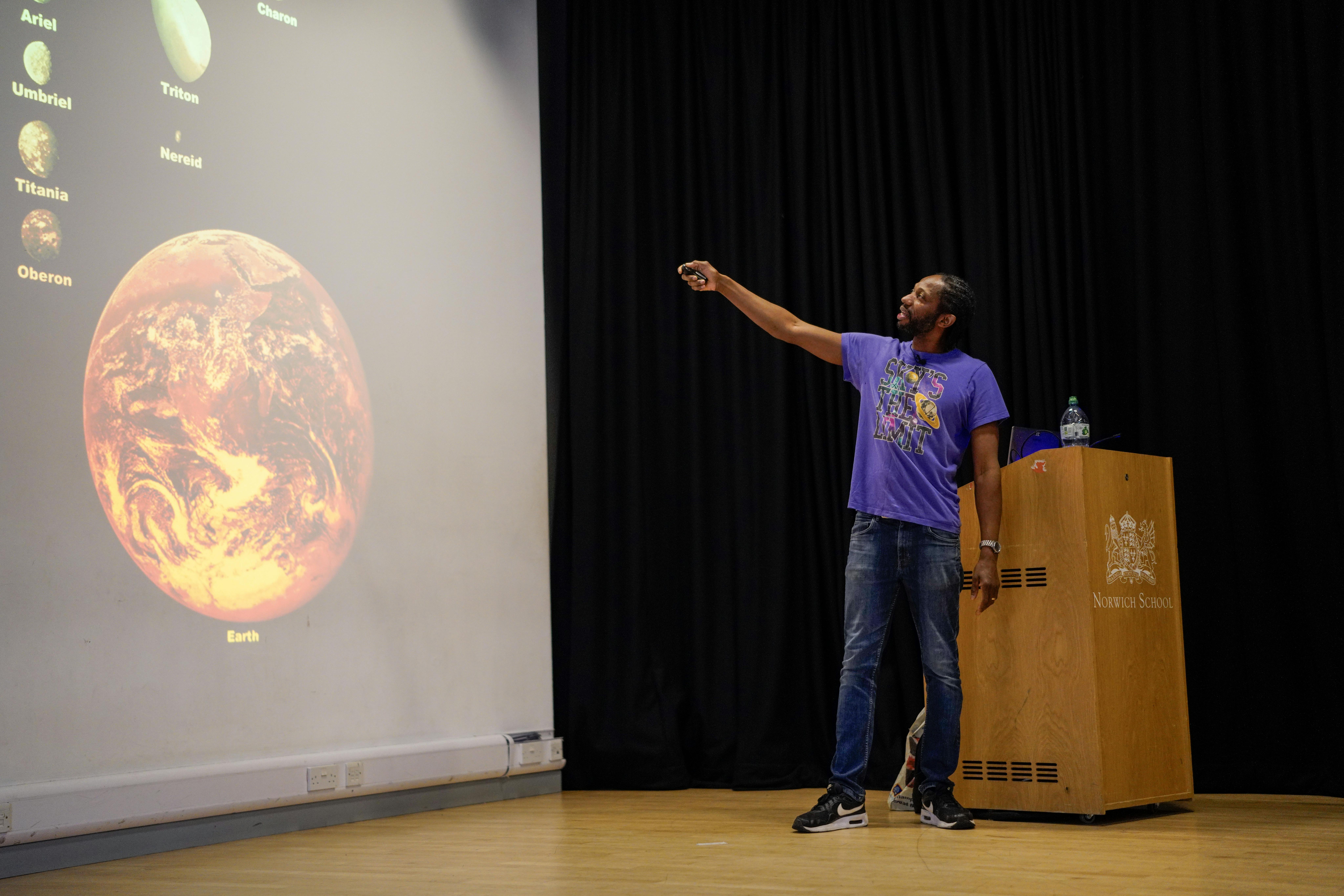A man on stage in a purple tshirt, with a slide show of planets on a large screen