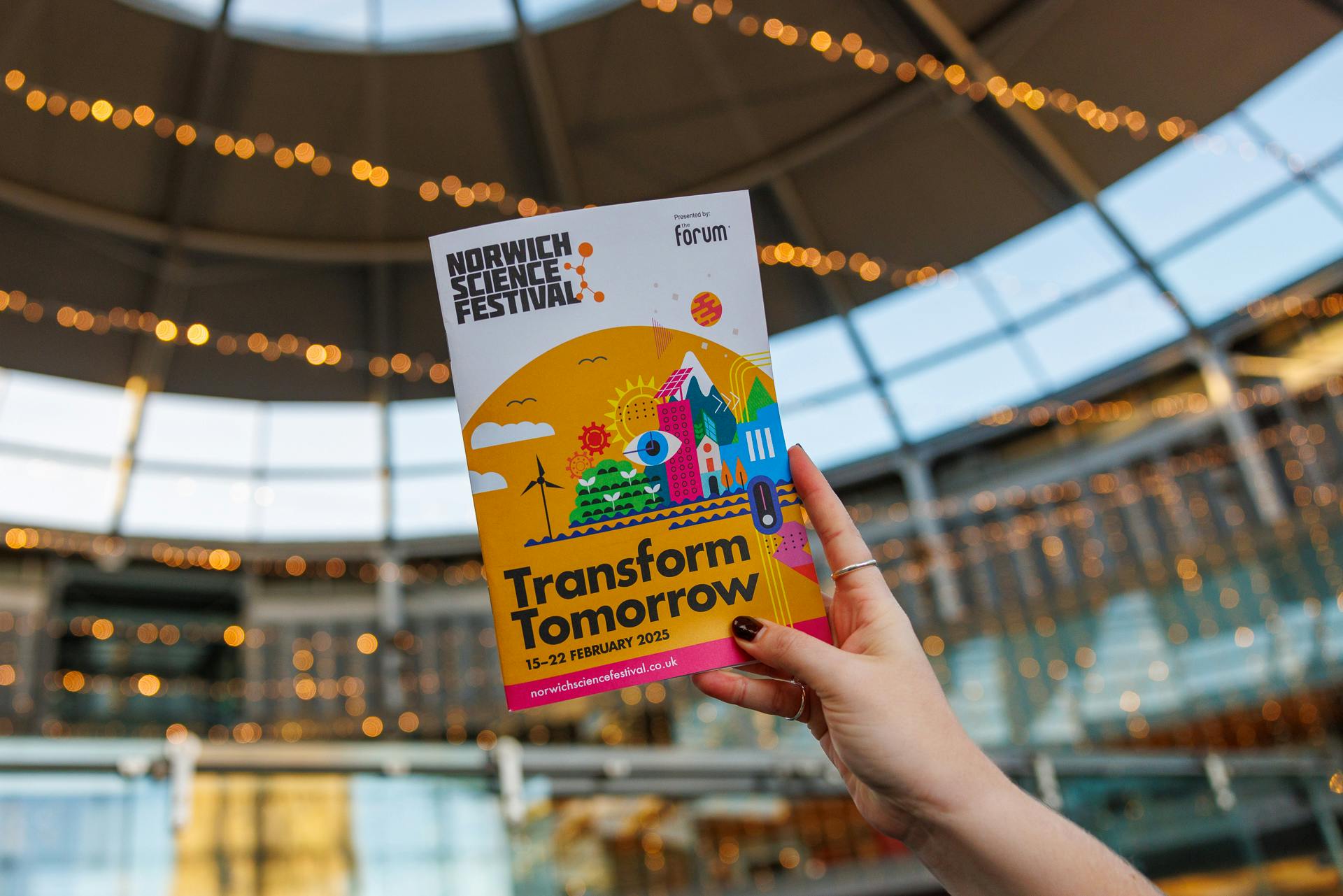 A hand holds up the 2025 Norwich Science Festival brochure with The Forum behind it