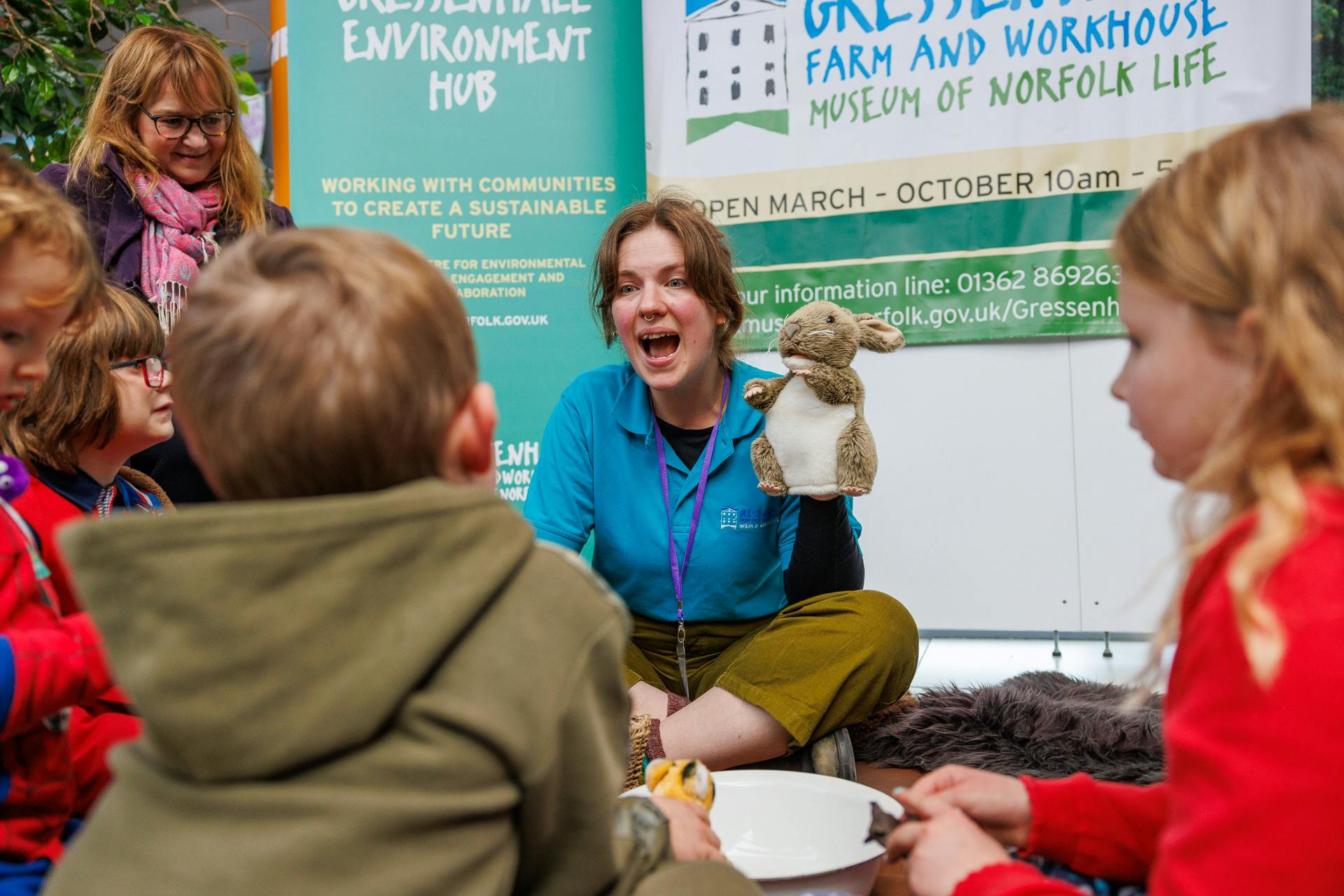 A woman in a blue polo shirt is speaking to a group of young children, holding a rabbit puppet.