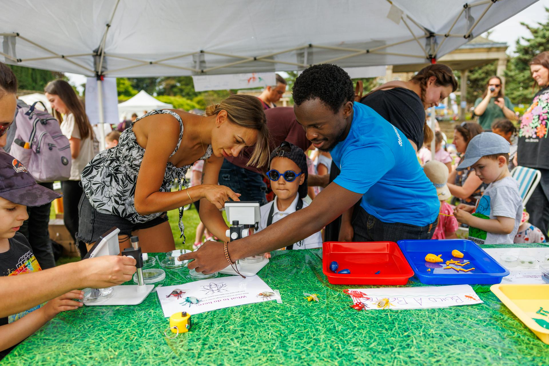 A Festival volunteer in a blue t-shirt helps a child and parent to look through a microscope