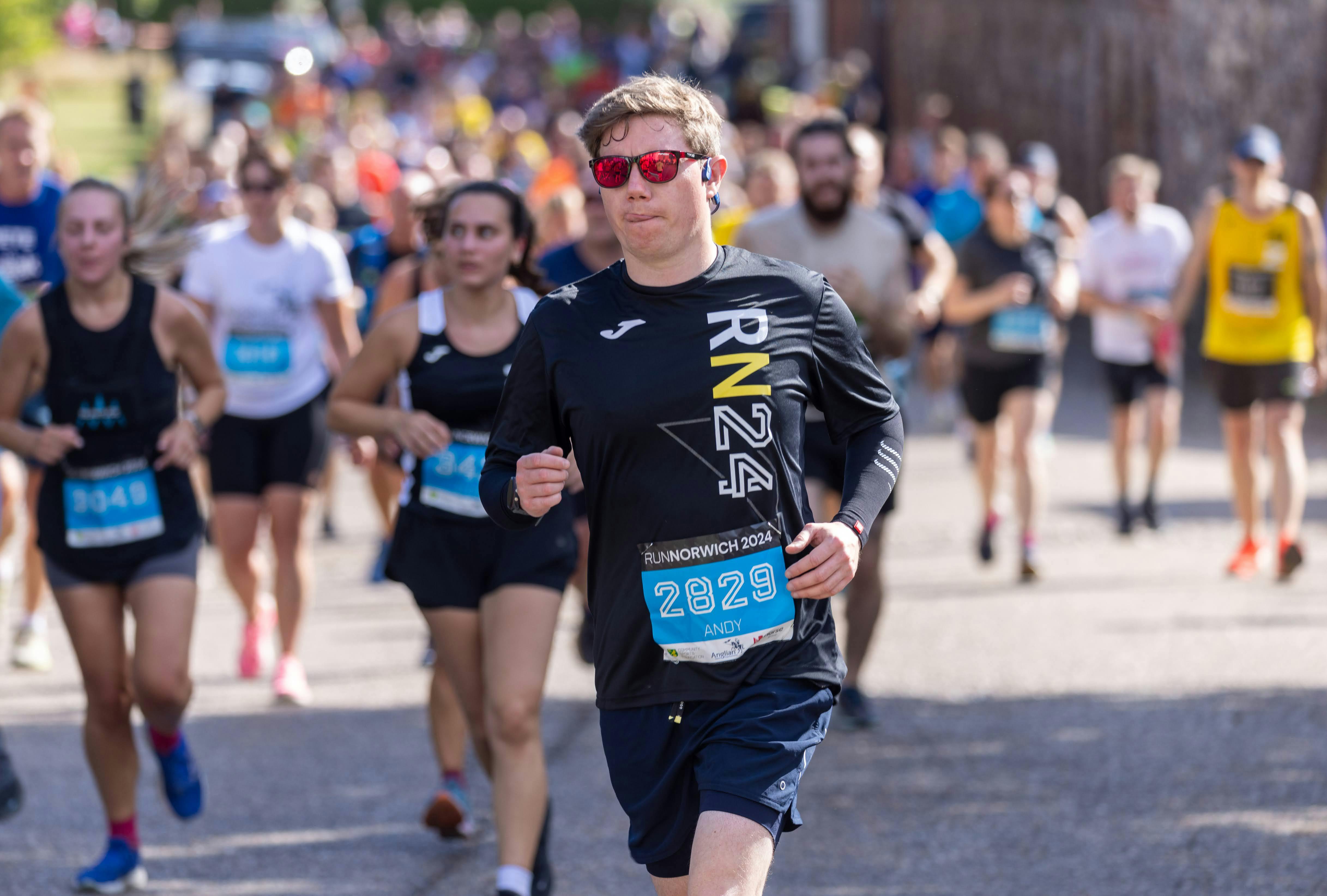 Man dressed in black t-shirt and shorts wearing red sunglasses concentrating while running.