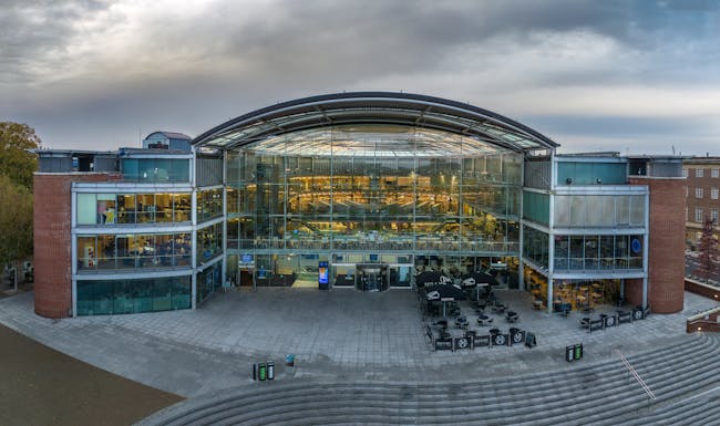 A drone has captured the front aspect of curved Forum building, with the warm glow of lights inside and a dynamic cloudy sky behind.
