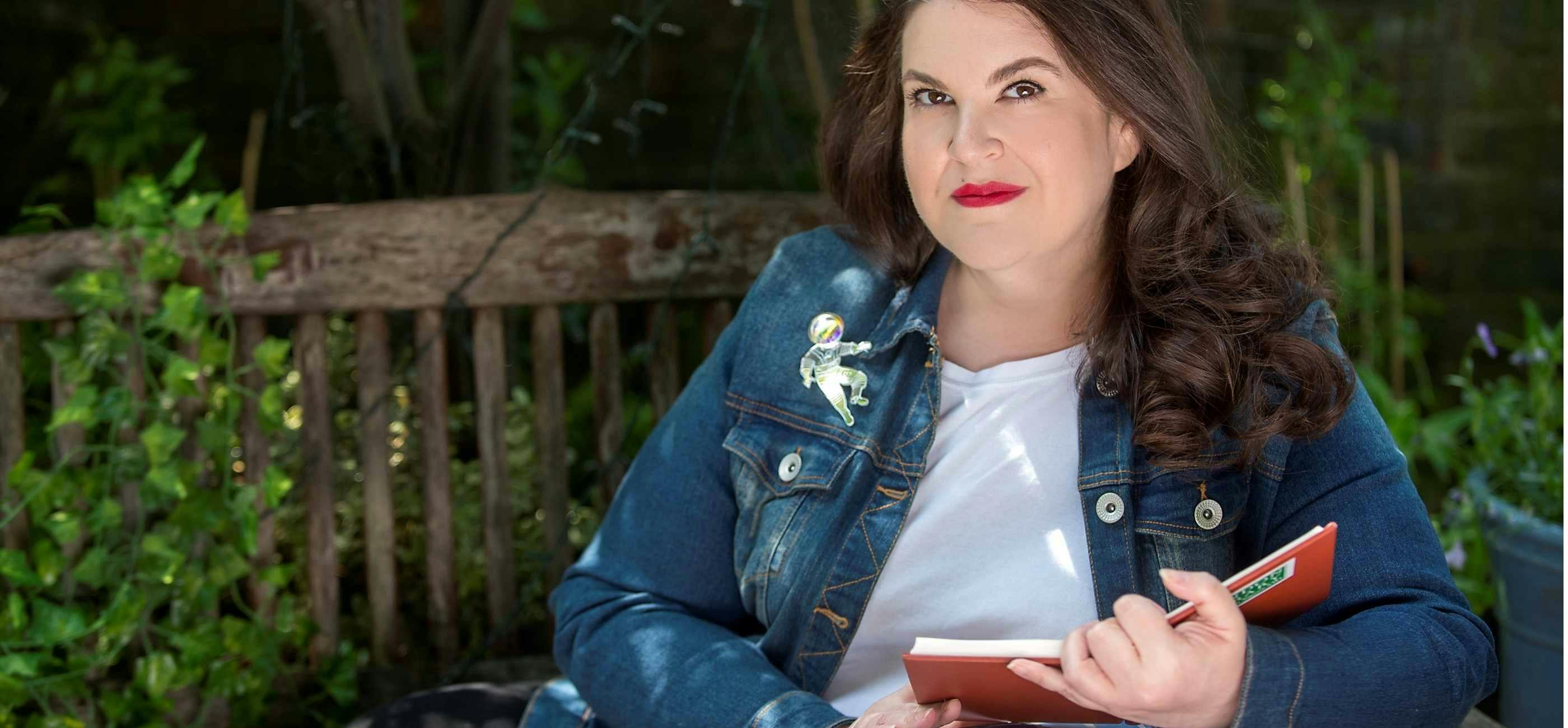 A photo of Naomi, a white woman with long dark hair, sitting on a bench with a book in her hands.
