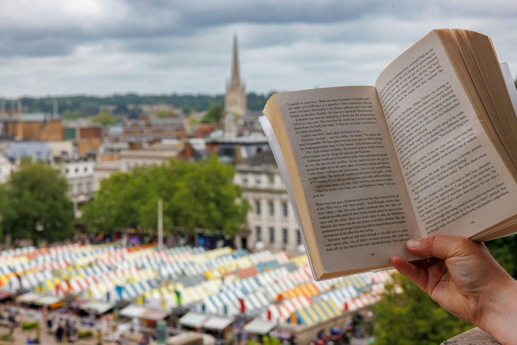 A hand holding a book against a backdrop of Norwich city centre - the market roofs and Norwich Cathedral